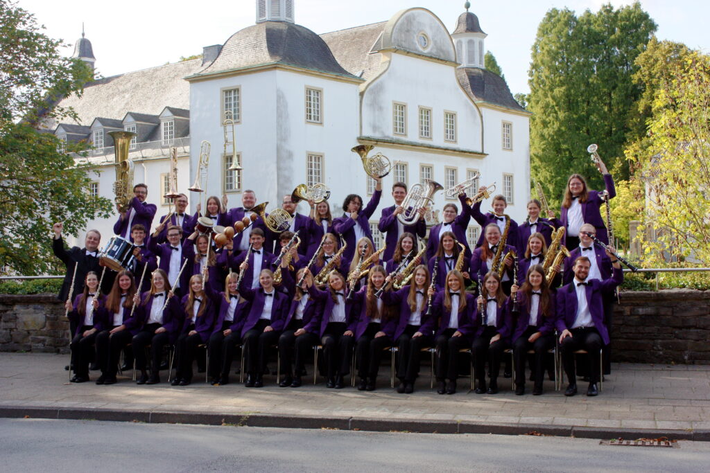 Das Schönebecker Jugend-Blasorchester in lila Uniformjacken vor Schloss Borbeck, die Musikerinnen und Musiker halten ihre Instrumente in die Höhe